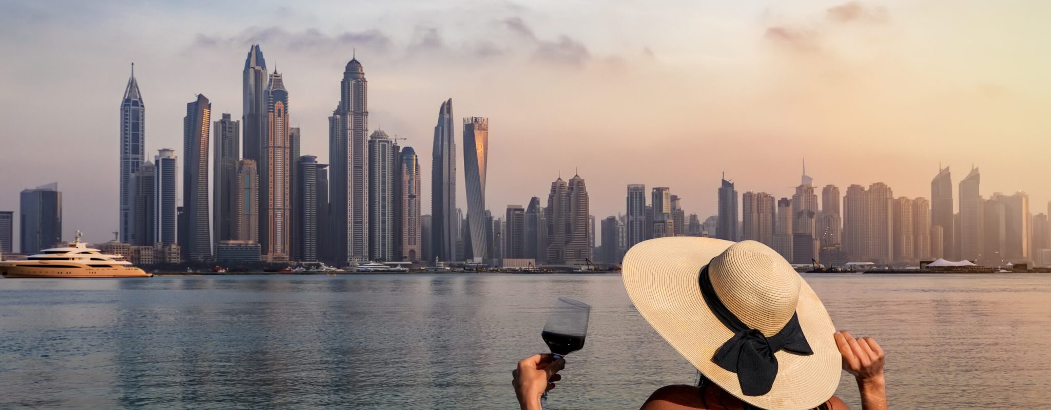 A elegant woman with a hat and a drink in her hand stands at the edge of a infinity pool and enjoys the view to the skyline of the Dubai Marina during sunset, UAE